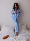 Woman in light blue pajamas holding a mug on a bed with white pillows and a croissant.